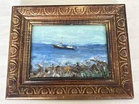 Framed oil painting depicting a boat on the ocean with rocky shore in front. Ornate gold-tone frame.