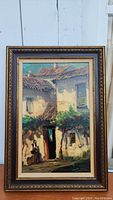 Framed oil painting showing village buildings, figure near doorway, tree, and textured surface.