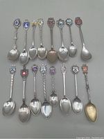 16 sterling silver souvenir spoons laid out showing enamelled crests and different handle designs.