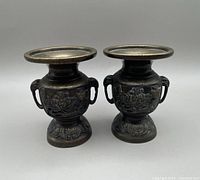 Photo showing two matching Japanese style bronze urns with wide rims, elephant head handles, floral relief designs, and leaf-pattern bases.
