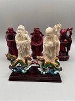 Five Chinese Fuk Luk Sau resin statues arranged in two rows. Two beige statues stand on colorfully painted dragon bases with one dragon tail missing. Three dark red lacquer statues stand separately in front.