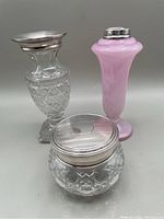 Group photo showing all three dresser items: clear cut glass cream jar with silver lid, pink opaque glass vase with silver lid, and clear cut glass vase with silver collar lid.
