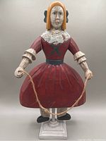 Front view of folk art wooden doll standing on a square base. Arms down holding a rope. Dressed in red with painted details.