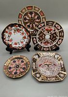 Five Imari style porcelain plates arranged, showing variety of shapes and intricate designs in blue, gold, red, and white.