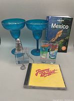 Lot items arranged: two blue plastic margarita glasses, two Cancun shot glasses, a Mexican sterling pendant on a clear cube with chain, Lonely Planet Mexico book, a CD case with 'Jimmy Buffett' on yellow cover.