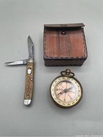 Compass, pocket tool and leather box arranged flat showing all items in lot