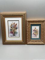 Two framed pressed flower art pieces by Kathryn Reith Blake displayed side by side. Larger one on left with gold frame and white mat; smaller on right with ornate gold frame and teal mat.