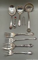Top-down view showing the nine silver plated serving pieces arranged flat. Includes 2 ladles, 1 large spoon, 4 serving forks, sugar tongs, and a teaspoon with amber glass finial.