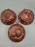 Top side view showing three copper molds with fruit designs of grapes, pomegranate, and strawberries, each with a hanging loop