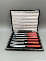 Set of six silver plated butter spreaders with salmon colored handles in a fitted box with padded lining.