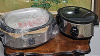Both items shown side by side on a wooden table against a floral wall painting. Slow cooker on right is black oval shape with glass lid, control dial visible. Roaster oven on left is larger, stainless steel with glass lid, partially wrapped in plastic.