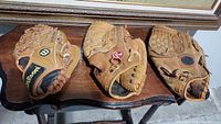 Three used baseball gloves laid out side by side on a dark wood surface. Left glove is Wilson brand, middle is Rawlings, right is unidentified darker glove with lattice web.
