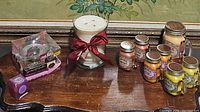 Various candles from Own Candle Company including mason jar style candles with metal lids labeled Pumpkin Cream Pie, Orange Creamsicle, Lemon Pound Cake, a large white candle wrapped with red bow, Glade Dewberry Dreams in packaging, and a Rose de Mai Blaze-On candle.