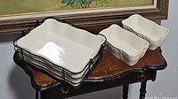 Three white ceramic casserole dishes with floral pattern shown on vintage wooden table. Large casserole fits in black metal basket with handle, two smaller loaf pans beside it.