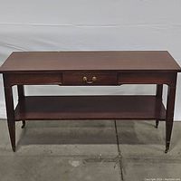 Front view of the Mersman console table showing the top, drawer with brass handle, lower shelf, and tapered legs with brass caps.
