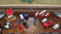 Overview of multiple small toy vehicles in emergency service colors including a red fire truck with ladder, a red rescue car, white utility vehicles, blue and black police vehicles, blue emergency personnel figures, and a small building model with a red roof.