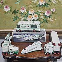 Photo of six Hess branded collectible toy vehicles on wooden table, showing toy truck with helicopter, space shuttle, dune buggy truck, car delivery truck, gasoline truck, and emergency ladder fire truck with some original boxes.