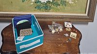 Photo of wooden table holding assorted jewelry items including bracelet, rings, earrings, brooches, coin/tissue holders, and a small storage bin with folded fabric items inside.