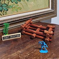 Photo showing Lincoln Logs assembled into a small log fort structure with a green plastic mini tree and a sign labeled 'FORT FRONTIER' on a wooden surface.