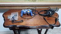 Three wired gaming accessories on a wooden table: blue Nintendo GameCube controller, black Logitech headset, and black Jabra earbud in plastic bag.