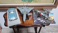 Photo showing a light blue Corvette model car on white base, a gray Tomcat plane on black stand, and a silver P-47 Thunderbolt plane on black base on a wooden table.