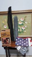 Photo showing boxed Coleman propane lantern, rolled chicken wire fence, black weed barrier fabric, folded American flag, and plastic bag with other items on a wooden table.