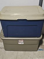 Blue plastic storage tote sitting on top of taupe Sterilite tote; both closed with lids.
