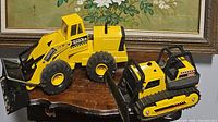 Photo showing both the yellow Tonka Mighty Diesel front loader and the metal bulldozer side by side on a polished wooden table with floral painting background.