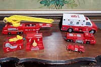Several large red toy emergency vehicles including a fire truck with a yellow ladder, an ambulance, and smaller fire trucks on a wooden surface.