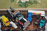 Front view of various toy trucks including the black Nylint tow truck with orange crane, yellow and green toy truck, smaller white and black trucks, blue semi truck with Hell-RuB trailer, red vintage style coin bank truck, and teal vintage style truck