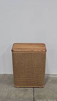 Front view of the wicker hamper showing rectangular shape, woven wicker exterior, and solid wood lid closed.