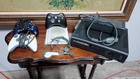 Xbox 360 console, power supply, two wired Xbox controllers (black and white), one wireless black controller, and headset shown on table.