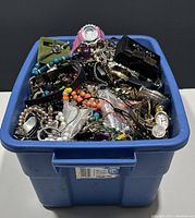 Blue plastic bin filled with tangled costume jewelry including watches, beads, chains, and bracelets
