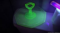 Glass cake plate with handle shown under UV light to reveal bright green fluorescence indicative of uranium content.