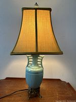 Front view of robin's egg blue pottery amphora shaped table lamp illuminated with beige fabric shade.