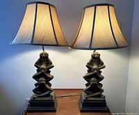 Pair of lit table lamps featuring stacked monkey figures representing the see no evil, hear no evil, speak no evil trio, in a dark bronze finish with beige fabric shades.