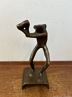 Front and angled side view of bronze frog bartender sculpture on wooden surface against white wall.