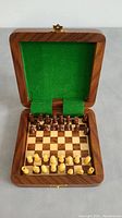 Open wooden case showing mini chessboard and full set of wooden chess pieces in starting position. Green felt lining inside lid.