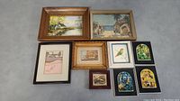 Lot of 9 small framed artworks arranged flat on grey carpeted floor showing front view.
