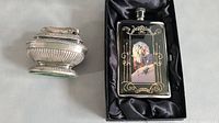 Photo showing the two items side by side: vintage silver-tone table lighter and black enamel flask with art deco style image.