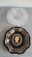 Photo showing both glass items from top view on grey surface: clear flower-shaped bowl and black decorative plate with gold detail.