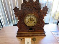 Wooden mantle clock with intricate floral carvings around the brass clock face. Visible soldering on the minute hand. Clock sits on a wooden table.