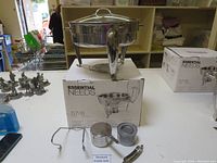 Photo of the stainless steel chafing dish on its box with accessories including fuel holders and tools placed in front on the table.