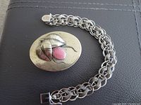 Silver tone charm bracelet alongside oval box with leaf motif and pink stone on dark background.