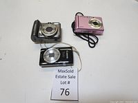 Overhead view of three compact digital cameras: silver Canon, pink Samsung, black Canon, arranged on table