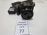 Front view of Panasonic Lumix DMC-FZ28 camera with lens cap on, showing the Lumix branding and attached battery charger in the background.