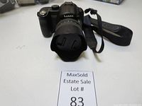 Front view of Panasonic Lumix camera with lens hood and strap, showing camera body and lens details.