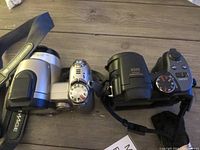 Top and front of silver Minolta and black GE digital cameras side by side on wooden surface