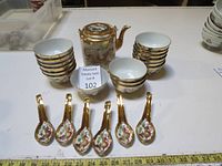 Photo showing the full set: one porcelain tea pot, 16 small tea bowls arranged in stacks, 6 tea spoons all decorated with gold trim and hand painted dragons.