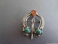 Front view of vintage Miracle brooch showing silver horseshoe shape with green stones on either side and orange stone on top center.